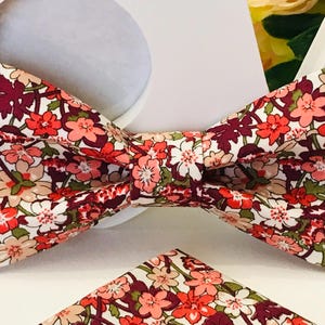 以下が含まれることがあります： 白い背景にピンク、赤、ボルドーの花が咲いた花柄の蝶ネクタイ。蝶ネクタイは布製で、クラシックな形をしています。