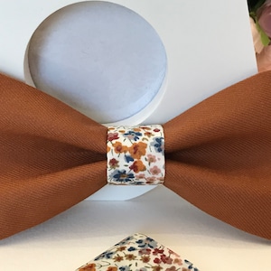 Puede incluir: Una pajarita marrón con un patrón floral blanco en la banda central. La pajarita está hecha de una tela texturizada.