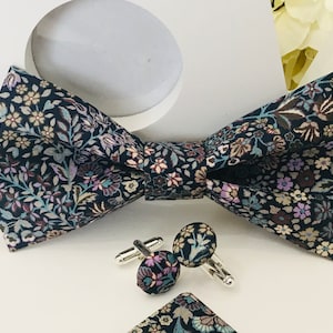 Può includere: Un papillon blu scuro con un motivo floreale in tonalità rosa, beige e turchese. Il papillon è abbinato a gemelli coordinati.