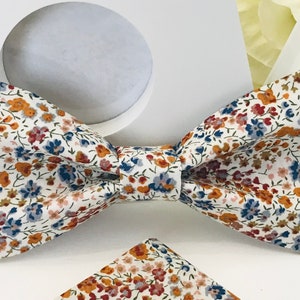 Può includere: Un papillon bianco con un motivo floreale in tonalità di blu, arancio e rosa. Il papillon è realizzato in un morbido tessuto di cotone e ha una forma classica a fiocco.