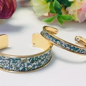 Può includere: Due braccialetti a polsino in metallo color oro. I braccialetti presentano un motivo floreale nei toni del verde, blu e giallo su sfondo bianco. I braccialetti sono aperti e hanno una finitura lucida. Lo sfondo include fiori.