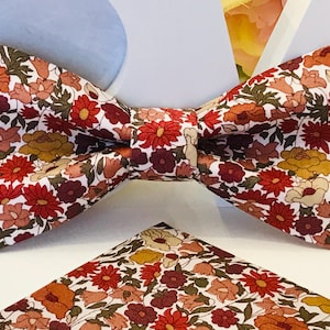 May include: A white bow tie with a floral pattern in shades of red, orange, and brown. The bow tie is made of cotton fabric and has a classic shape.