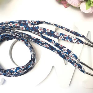 May include: Navy blue shoelaces with a floral pattern of white flowers and blue leaves. The shoelaces have silver metal aglets. The laces are arranged in a decorative pattern on a white surface.