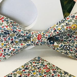 Può includere: Un papillon bianco con un motivo floreale in tonalità di rosso, blu, giallo e verde. Il papillon è abbinato a un fazzoletto da taschino.