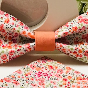 Peut inclure: Un ensemble de nœud papillon et de pochette assortie à motifs floraux. Le nœud papillon présente un fond blanc avec un motif de petites fleurs roses, oranges et vertes. La bande centrale est de couleur pêche unie. La pochette a le même motif floral.