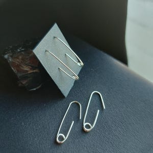 Op de afbeelding: Zilverkleurige veiligheidsspeld oorbellen. Eén paar wordt getoond op een zwarte vierkante kaart en een ander paar ligt op een donkere ondergrond. De oorbellen hebben een eenvoudig, minimalistisch ontwerp.
