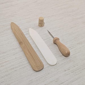 May include: A collection of bookbinding tools, including a bamboo folder, a bone folder, a cork, and an awl with a wooden handle. The tools are arranged on a light-colored surface.