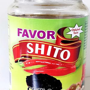Op de afbeelding: Een glazen pot Favor Shito chilisaus met een gouden metalen deksel. Het etiket is lichtgroen met een rode lintafbeelding, de tekst "FAVOR SHITO", twee rode pepers en een "Original Taste"-ster. De onderste tekst luidt "Sauce Chili".