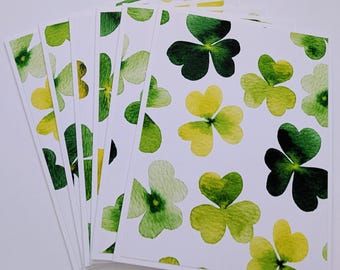 May include: A stack of white cards featuring a watercolor pattern of green and yellow shamrocks. The shamrocks vary in shades of green and yellow, with some appearing darker and others lighter. The cards are arranged in a fan shape.