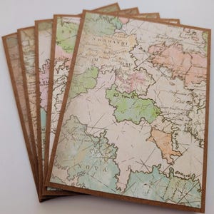 May include: A set of folded greeting cards featuring a vintage-style map print on the front. The map displays pastel-colored regions in shades of green, pink, and tan, overlaid with black text and thin intersecting lines on a brown cardstock base.