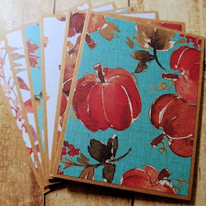 May include: A stack of greeting cards with various autumn-themed designs. The cards feature images of pumpkins, leaves, and floral patterns in shades of red, orange, and teal. Each card has a brown border.
