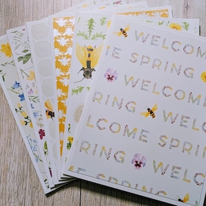 May include: A collection of decorative paper sheets with floral and bee designs. One sheet reads "WELCOME SPRING" in a floral font. Other sheets feature patterns of flowers, bees, and honeycomb. The color palette includes yellow, white, and green.