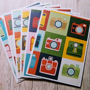 May include: A collection of colorful, patterned cards featuring vintage camera illustrations. Each card showcases a different camera design in a square grid, with a variety of colors including red, green, yellow, and blue. The cards are arranged on a light wood surface.