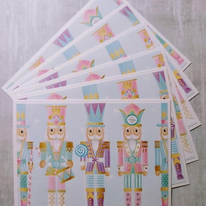 May include: A set of six pastel blue and pink Christmas cards featuring a row of five nutcracker soldiers with candy cane accents. The nutcrackers are wearing colorful hats and uniforms with gold trim. The background is a light blue with snowflakes.