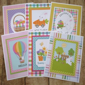 May include: Set of six Easter-themed greeting cards featuring colorful checkered and striped patterns. The cards feature bunnies, a hot air balloon, a garden scene, and a basket of eggs. The text on the cards includes "Easter's On", "You Make", "Hello!" and "Thanks".