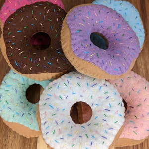 May include: Five felt donuts with colorful sprinkles. The donuts are in a stack with the pink donut on top and the white donut on the bottom. The donuts are brown on the bottom and have a hole in the center.