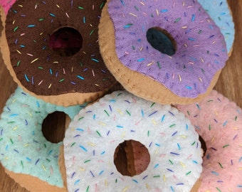May include: Five felt donuts with colorful sprinkles. The donuts are in a stack with the pink donut on top and the white donut on the bottom. The donuts are brown on the bottom and have a hole in the center.