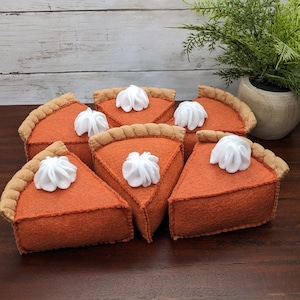 May include: Six felt pumpkin pie slices with white whipped cream topping. The slices are arranged in a circle on a brown wooden surface.