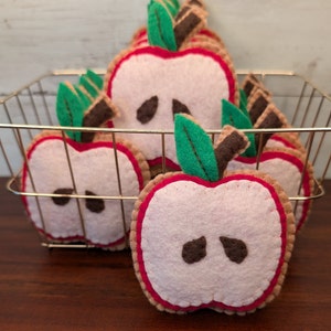 May include: A basket filled with felt apple ornaments. The apples are red with a pink center and brown seeds. Each apple has a green felt leaf and a brown felt stem.