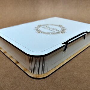 May include: A white wooden box shaped like a book, with a decorative gold emblem and the name "Natalia" and "#15 Anos" printed on the top. The box has black edges and a light brown base, suggesting a keepsake or gift box.