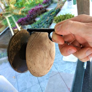 Könnte beinhalten: Ein Flaschenverschluss aus Holz und Metall mit einem schwarzen Griff und einer kleinen Kette. Der Verschluss hat einen hellbraunen, oval geformten Körper und eine dunkelbraune, kreisförmige Oberseite. Der Griff wird von einer Hand gehalten.