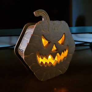 Pode incluir: Uma lanterna de abóbora de Halloween de madeira com um rosto esculpido e um caule. A abóbora é marrom escuro com uma luz laranja brilhante no interior. A decoração está em uma superfície escura.