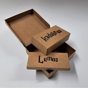 May include: Cardboard boxes, one open, with the words "Kadabrus" and "Lumos" printed in black gothic font. The boxes are brown and appear to be empty, with a white background.