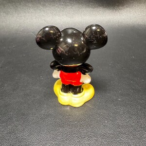 May include: A small, glossy figurine of Mickey Mouse, viewed from the back. The figure has large, black ears, a red jacket, white gloves, and black shoes. It stands on a yellow base, set against a dark background.