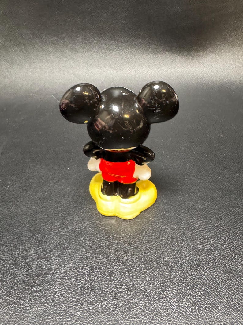 May include: A small, glossy figurine of Mickey Mouse, viewed from the back. The figure has large, black ears, a red jacket, white gloves, and black shoes. It stands on a yellow base, set against a dark background.