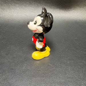 May include: A ceramic figurine of Mickey Mouse, featuring a black head and ears, a white face, and a red shirt. The figure has yellow shoes and white gloves. The figurine is a collectible item.