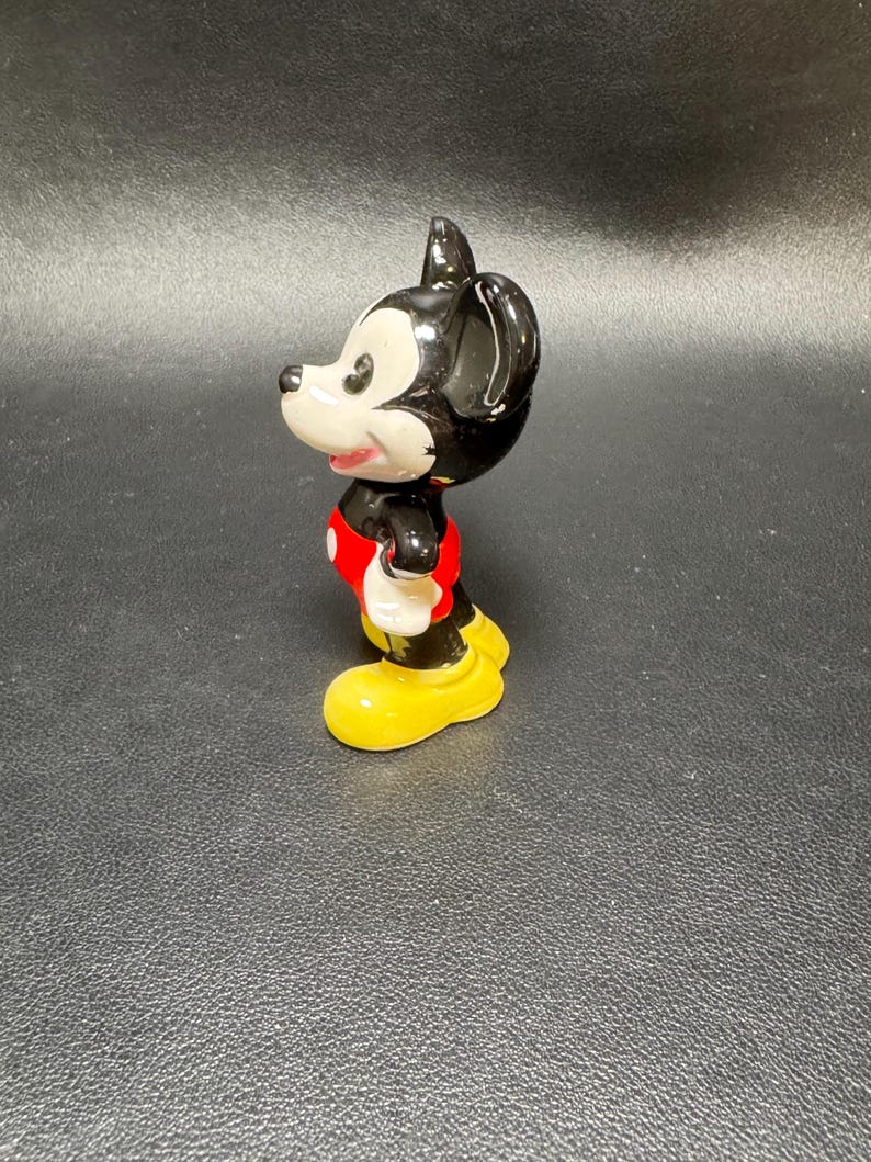 May include: A ceramic figurine of Mickey Mouse, featuring a black head and ears, a white face, and a red shirt. The figure has yellow shoes and white gloves. The figurine is a collectible item.