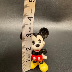 May include: A small, vintage Mickey Mouse figurine, approximately 2 inches tall, with black ears, a white face, and red shorts with white buttons. The figurine has yellow shoes and a black body. A measuring tape is next to the figurine.
