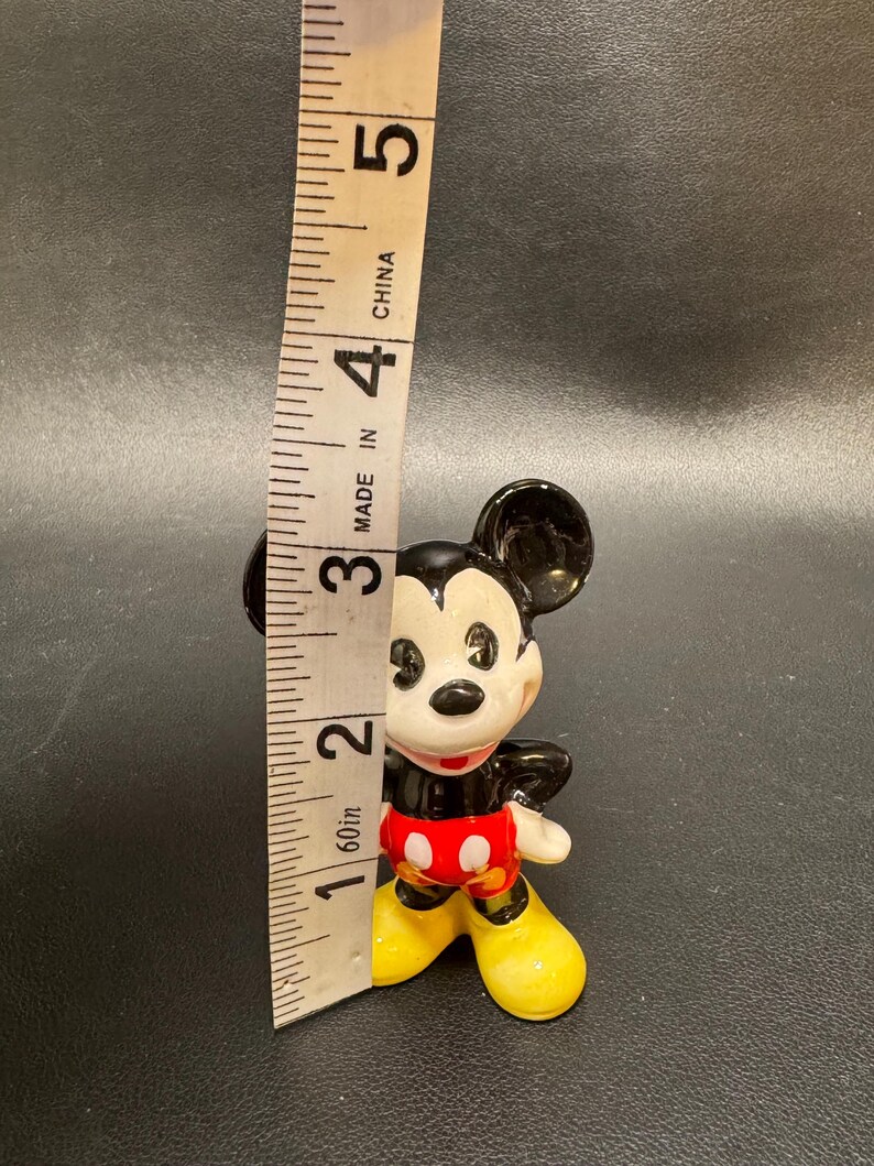 May include: A small, vintage Mickey Mouse figurine, approximately 2 inches tall, with black ears, a white face, and red shorts with white buttons. The figurine has yellow shoes and a black body. A measuring tape is next to the figurine.