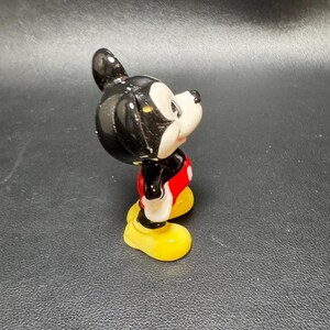 May include: A ceramic figurine of Mickey Mouse, featuring a black head and ears, a white face, red shorts, yellow shoes, and black arms. The figure is posed in a side view, showcasing its classic design and vibrant colors.