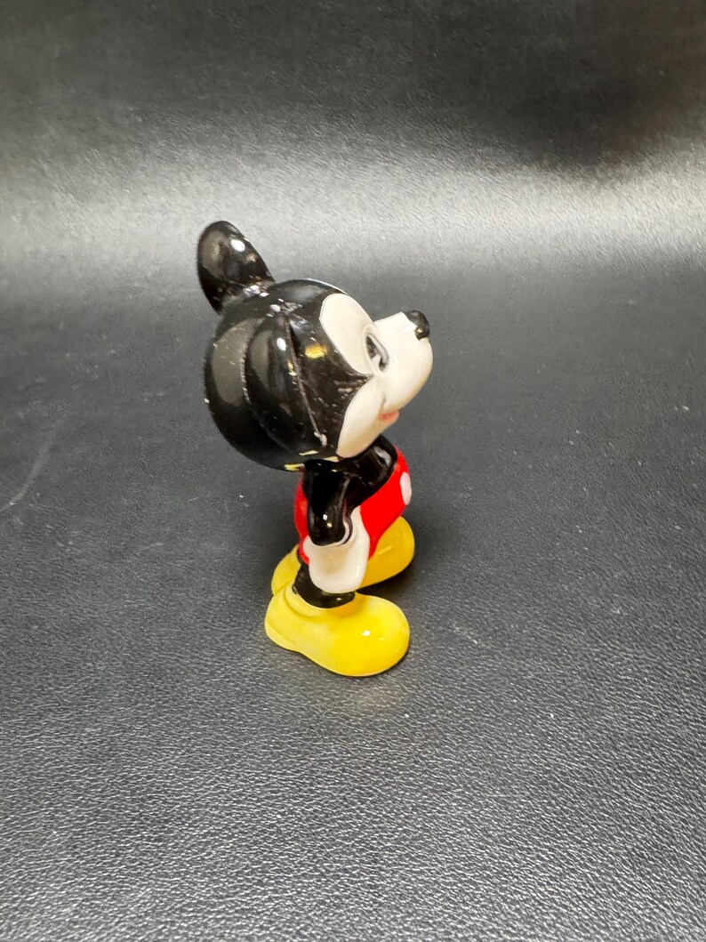May include: A ceramic figurine of Mickey Mouse, featuring a black head and ears, a white face, red shorts, yellow shoes, and black arms. The figure is posed in a side view, showcasing its classic design and vibrant colors.