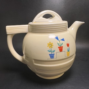 May include: A cream-colored ceramic teapot featuring a horizontal ribbed design on the body and handle. The side is decorated with a colorful graphic of three potted flowers in blue, orange, and yellow pots arranged on a stepped shelf.
