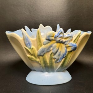 May include: A ceramic bowl featuring a scalloped rim and a raised relief design of blue and yellow iris flowers with green stems. The bowl has a cream-colored body with a light blue base and interior, showcasing a hand-painted floral motif.