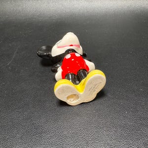 May include: A ceramic Mickey Mouse figurine, lying on its back. The figure features a white face, black ears, and a red shorts. The shoes are yellow, and the figure has a black body and a white collar.