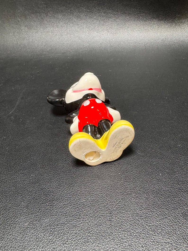 May include: A ceramic Mickey Mouse figurine, lying on its back. The figure features a white face, black ears, and a red shorts. The shoes are yellow, and the figure has a black body and a white collar.