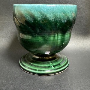 May include: A green ceramic bowl with a pedestal base. The bowl has a glossy finish with shades of dark and light green. The base is a lighter green with concentric circles. The bowl is likely vintage.
