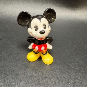 May include: A ceramic figurine of Mickey Mouse. The figure has black ears, a black body, white gloves, red shorts, and yellow shoes. The figurine is standing upright with its arms at its sides.