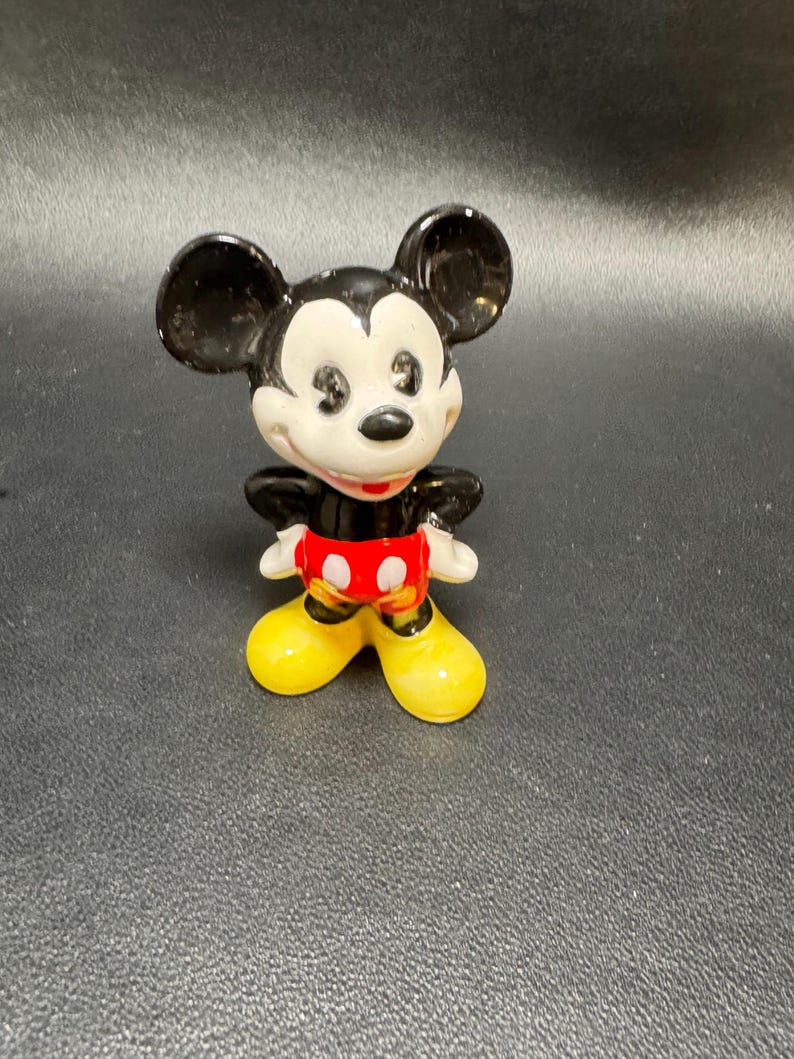 May include: A ceramic figurine of Mickey Mouse. The figure has black ears, a black body, white gloves, red shorts, and yellow shoes. The figurine is standing upright with its arms at its sides.