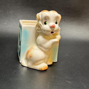 May include: A vintage ceramic planter featuring a white puppy with orange and tan accents. The puppy is holding onto a rectangular planter with a light blue background. The planter has a square opening and is likely for small plants or flowers.
