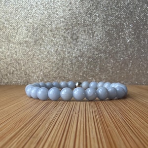 May include: A stretch bracelet composed of smooth, round, light blue beads. The beads have a glossy finish and are uniform in size. A single small, metallic silver-toned bead is positioned near the centre of the circular arrangement of light blue stones.