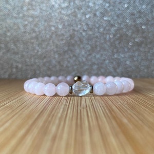 May include: A pink gemstone bracelet with a clear crystal bead in the centre. The bracelet is on a light brown wooden surface.