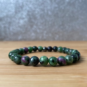 May include: A bracelet made of polished, round gemstones in shades of green, red, and black. The beads are strung together to form a circular shape. The bracelet is presented on a wooden surface.