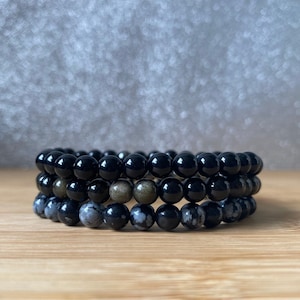 K&ouml;nnte beinhalten: Drei &uuml;bereinanderliegende Armb&auml;nder aus schwarzen, grauen und goldenen Perlen. Die Armb&auml;nder sind aus nat&uuml;rlichen Steinen.