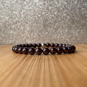 May include: A dark red gemstone bracelet with a silver clasp. The bracelet is made of round beads and is lying on a wooden surface.