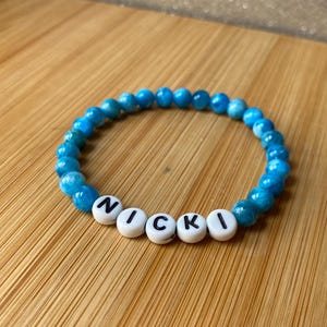 May include: A bracelet featuring blue gemstone beads and white letter beads spelling "NICKI". The beads are strung on an elastic band. The bracelet is displayed on a wooden surface with a natural grain pattern.