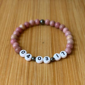 May include: A beaded bracelet featuring round, dusty rose-coloured stone beads interspersed with small, dark metallic spheres. The centre section includes white, cylindrical plastic beads printed with the black numbers 04, 03, and 17.